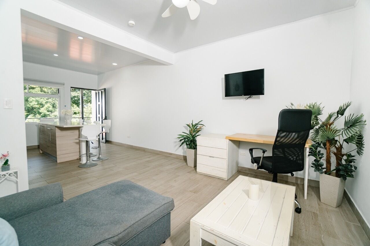 A modern living room with a grey sofa, a white coffee table, a black office chair, a wooden desk, and a TV mounted on the wall. There are two potted plants and a kitchen area visible in the background.
