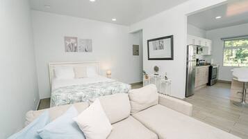 A modern, well-lit bedroom with a large bed, a sofa, and a kitchenette in the adjacent room.