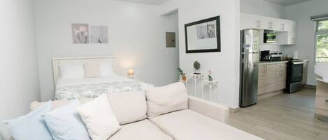 A modern, well-lit bedroom with a large bed, a sofa, and a kitchenette in the background.