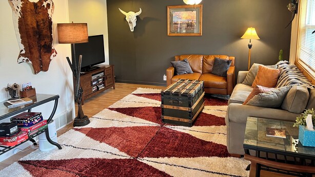Un salon avec un plafond en bois, un téléviseur fixé au mur, un canapé en cuir, une table basse en verre et un tapis à motifs.