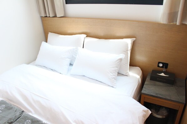 A neatly made bed with white linens and pillows, a wooden headboard, a small bedside table with a tissue box, and a power outlet. - ION HOTEL DONGDAEMOON (Seoul)