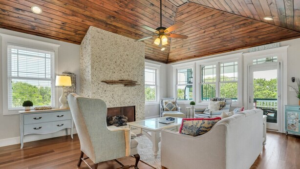 A spacious living room with a fireplace, wooden ceiling, and large windows offering a view of greenery. - Tybee Skies: 4 BDR 4 Bath Oceanfront Home w Pool (Tybee Island)