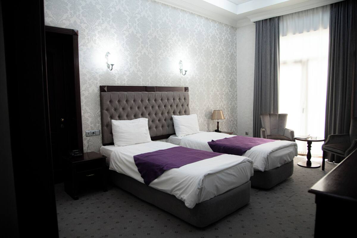 A hotel room with two beds, a nightstand, a lamp, a chair, and a desk.