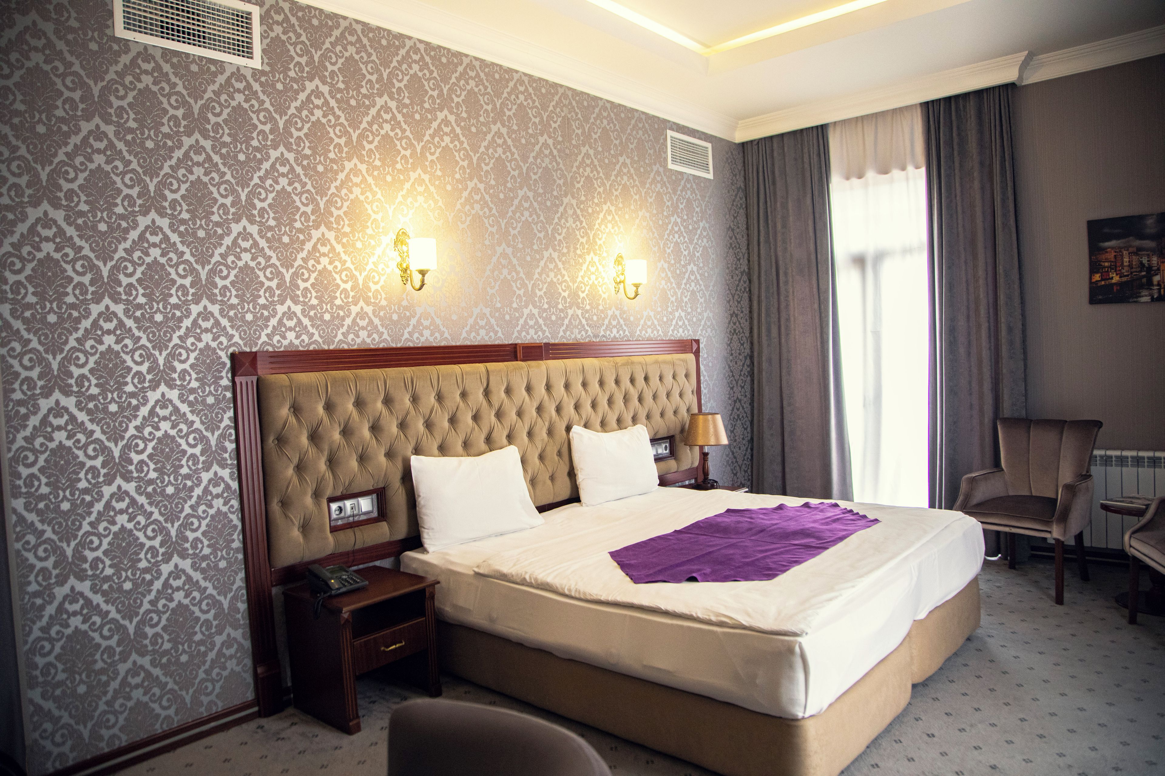 A hotel room with a large bed, two bedside lamps, a nightstand, a chair, and a window with curtains.