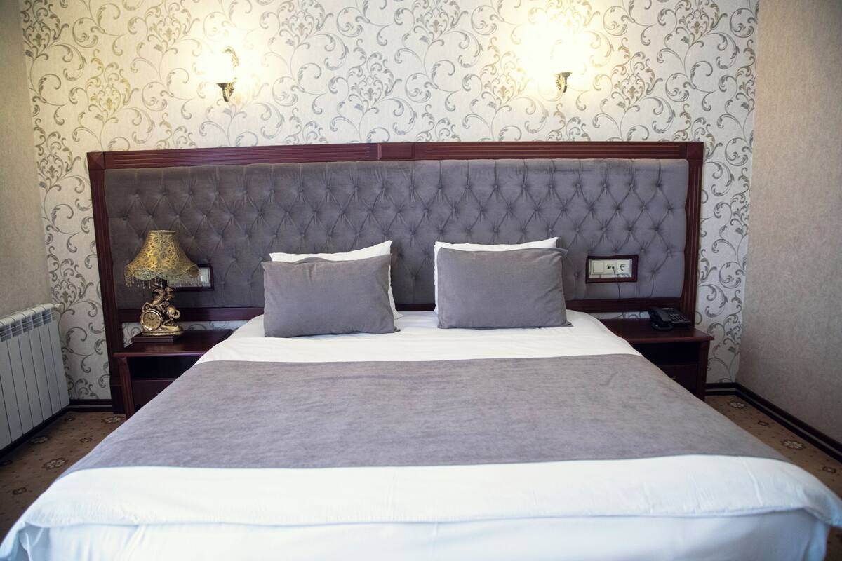 A neatly made bed with a grey headboard and two pillows, a bedside lamp, and a telephone on a wooden nightstand.