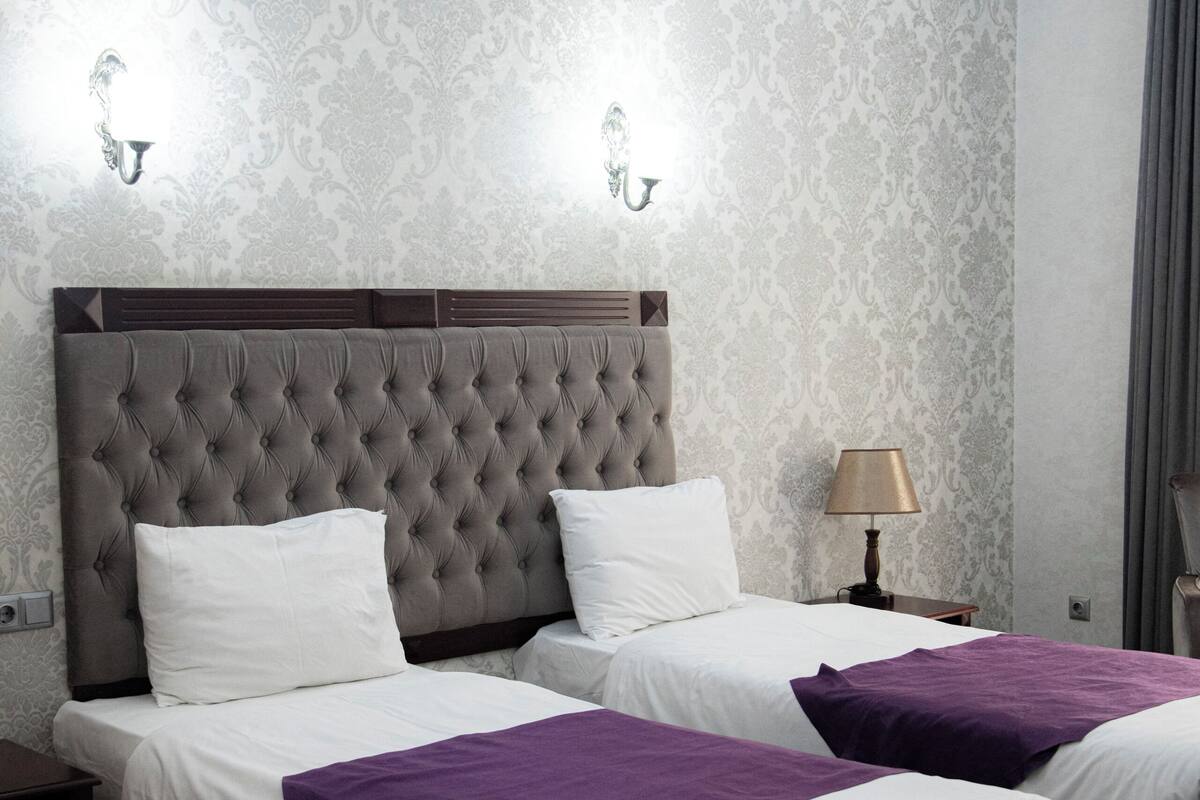 A double bed with a tufted headboard, two bedside tables, a lamp, and a patterned wallpaper.