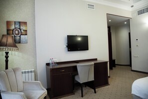 A hotel room with a desk, a television, a lamp, a chair, and a radiator.