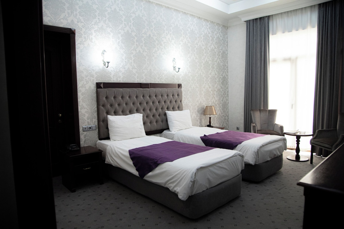 A hotel room with two beds, a nightstand, a lamp, a chair, and a desk.
