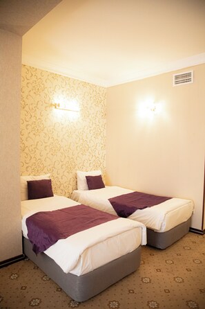 Una habitación de hotel con dos camas individuales, cada una con una colcha y almohada moradas, un papel tapiz estampado y un respiradero en el techo.