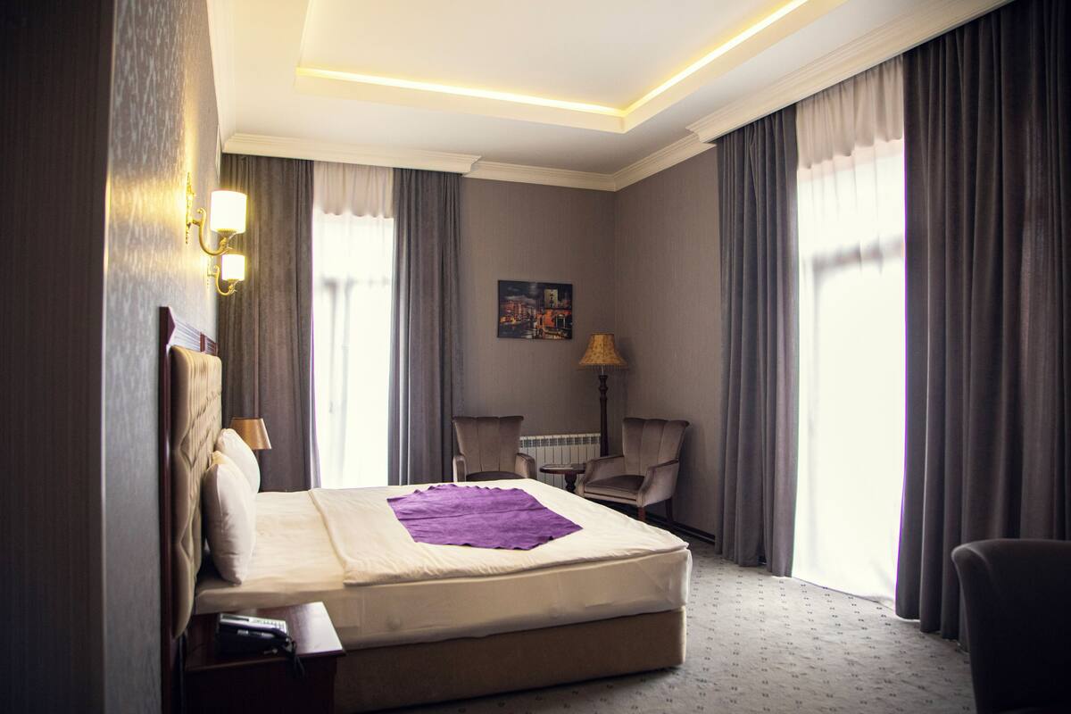 A hotel room with a bed, two armchairs, a lamp, a painting on the wall, and a window with curtains.