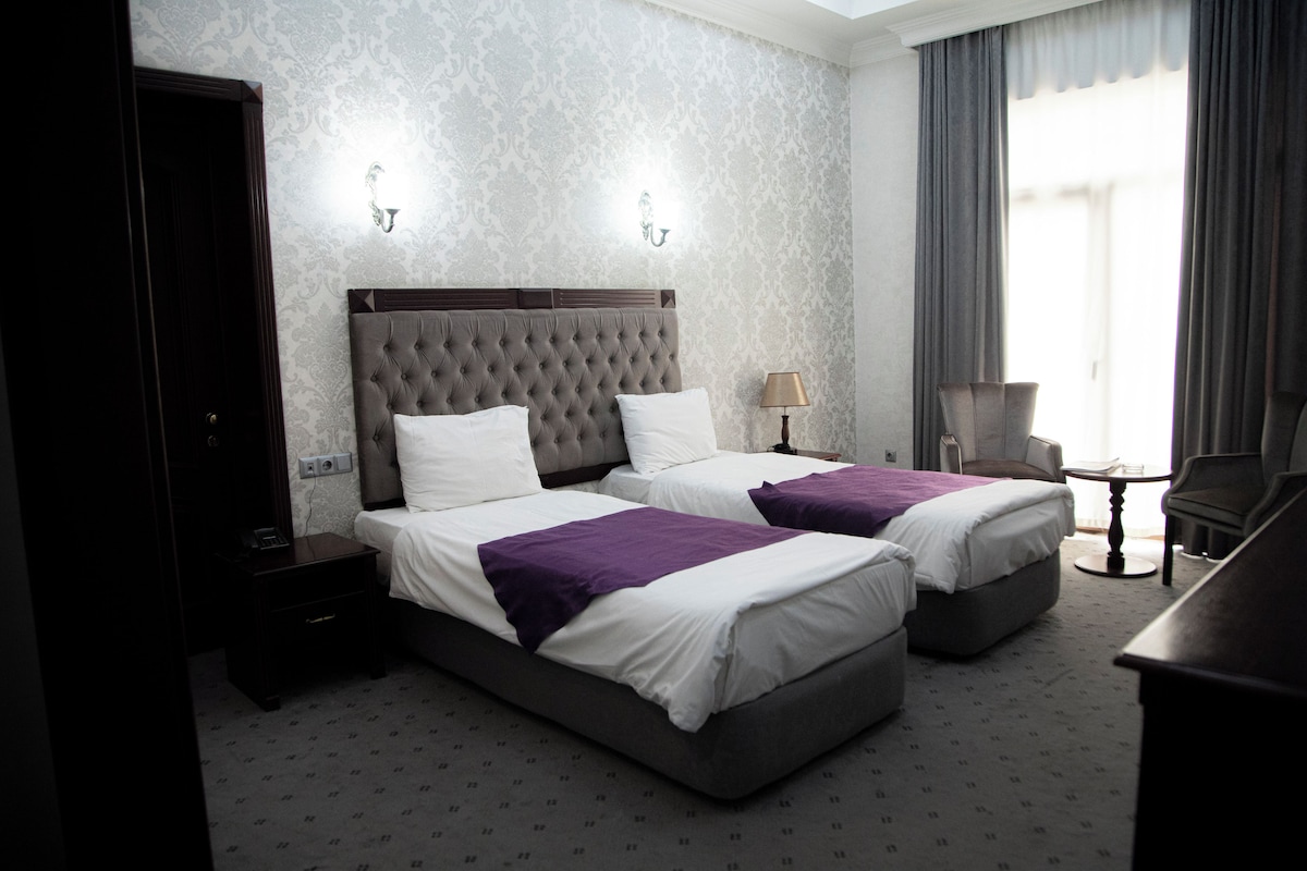 A hotel room with two beds, a nightstand, a lamp, a chair, and a small table.