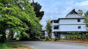 A two-story building with a grey roof and white walls, surrounded by lush greenery and a paved road. - Hilo Lakefront Studio, Pool, Walk to Downtown (Hilo)