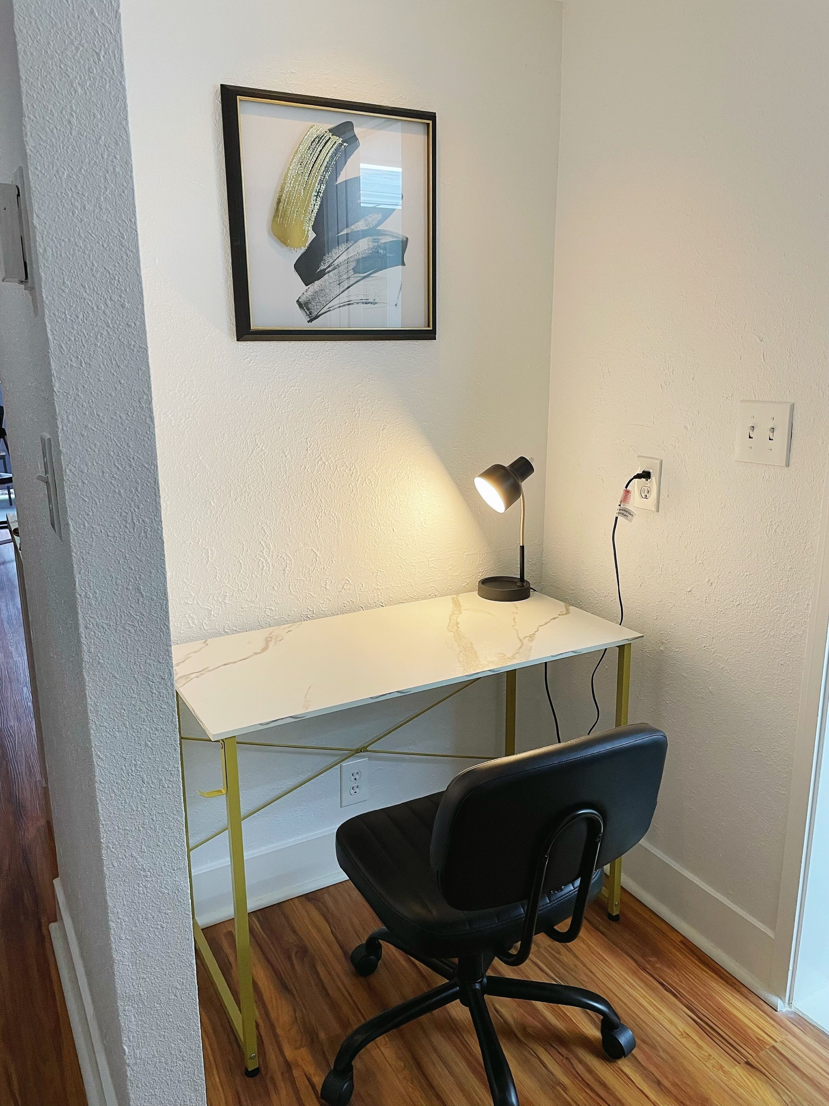 Ein kleiner, moderner Arbeitsraum mit einem weißen Marmor-Schreibtisch, einem schwarzen Bürostuhl, einem gerahmten abstrakten Gemälde an der Wand und einer Schreibtischlampe.