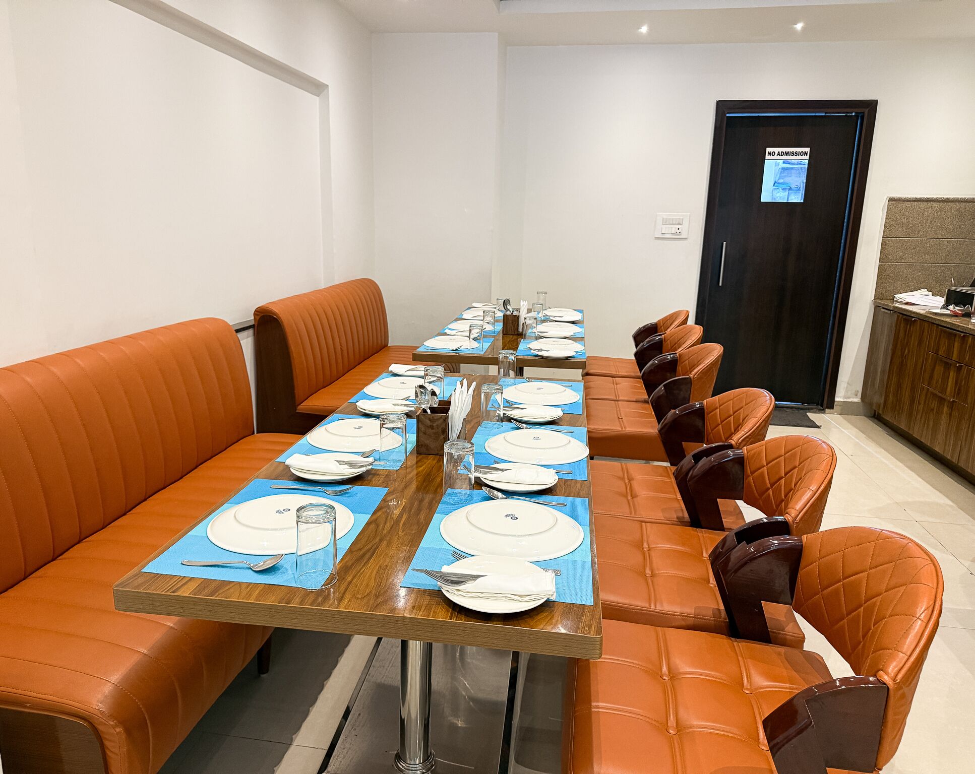 A dining area with a long table set for multiple people, orange cushioned benches, and a door with a 'No Admission' sign.
