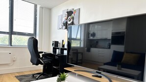 Un salon moderne avec un grand téléviseur à écran plat, un fauteuil de bureau noir, une table basse blanche agrémentée d’une plante, et un coin cuisine en arrière-plan.