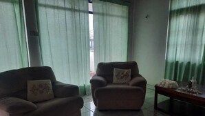 Ein Zimmer mit zwei Sesseln, einem kleinen Tisch und einem Fenster mit grünen Vorhängen.