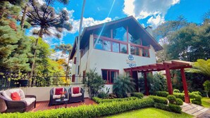 A two-story building with a sign reading "Pousada do Lago". There are wicker chairs with red cushions on a wooden deck, surrounded by well-maintained hedges and trees. - Pousada do Lago Gramado (Gramado)
