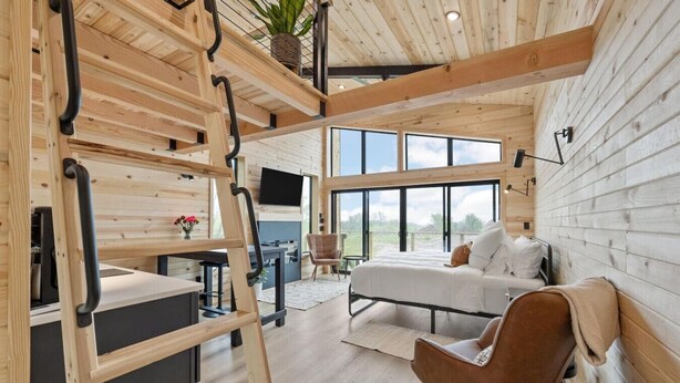 A modern log cabin interior with a loft bed, a desk, and a large window offering a view of the outdoors.