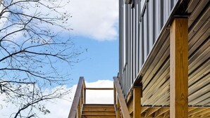 A wooden staircase leading to a building with a metal railing, a welcome mat at the bottom, and a clear sky in the background.