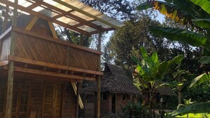 Un edificio rústico de madera con techo de paja, un porche cubierto y un pequeño jardín con plantas de plátano.