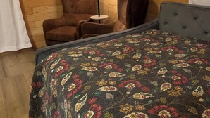 Une pièce chaleureuse aux murs en bois, avec un lit recouvert d’une courtepointe à motifs floraux, un fauteuil en cuir marron et un lampadaire.
