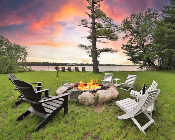 Un hogar de fuego rodeado de sillas Adirondack, un espacio con césped y vistas al lago.