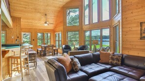 A spacious wooden cabin interior with a large leather sofa, dining area, and multiple windows offering views of the outdoors.