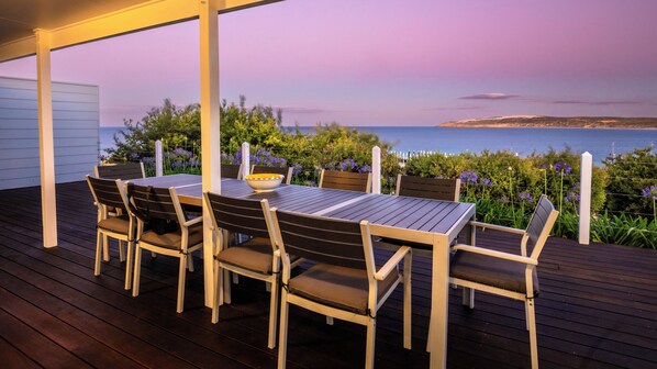 A covered outdoor dining area with a table and chairs, overlooking a body of water and hills.