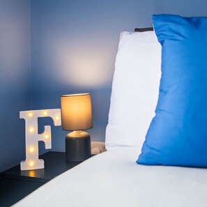 A neatly made bed with a blue pillow and a white pillow, a nightstand with a lamp and illuminated letter 'F', and a wall with a blue hue.