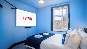 A room with a bed, a window with bars, a wall-mounted screen displaying "NETFLIX", and a ceiling light fixture.