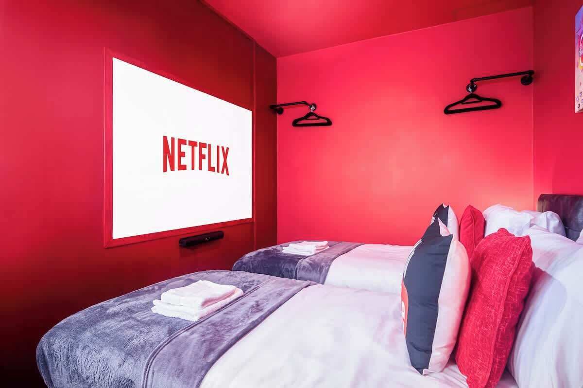 Een kamer met twee bedden, rode wanden, een groot raam met het Netflix logo, en twee zwarte kledinghangers aan de muur.