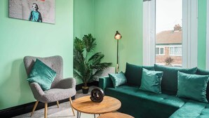 Una habitación con una pared verde, un sillón gris, un sofá verde, una mesita redonda pequeña y una lámpara de pie.