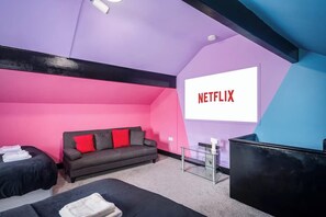 A room with a pink and black color scheme, a gray sofa with red cushions, a bed with a black headboard, and a large screen displaying the Netflix logo. - Social Media Adventure - 3 Bed 10% off Long stays (Doncaster)