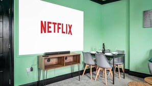 Una habitación con una pantalla grande mostrando el logotipo de Netflix, un escritorio de madera con una computadora, dos sillas y una mesa de comedor con copas de vino.