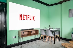 Une pièce dotée d’un grand écran affichant le logo de Netflix, d’un bureau en bois avec un ordinateur, de deux chaises et d’une table à manger avec des verres à vin.