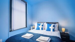 Un dormitorio con cama, ropa de cama azul, una mesita de noche con lámpara y una ventana con persianas.
