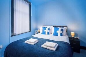 Un dormitorio con cama, ropa de cama azul, una mesita de noche con lámpara y una ventana con persianas.