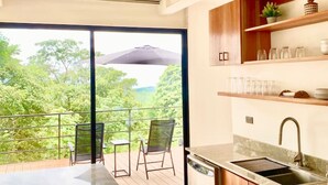 A modern kitchen with a large window, a balcony with chairs, and a view of greenery.