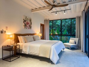 A modern bedroom with a large bed, bedside lamps, a ceiling fan, and a view of greenery through large windows.