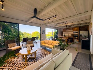 A spacious living area with a ceiling fan, large windows, and a kitchen in the background.