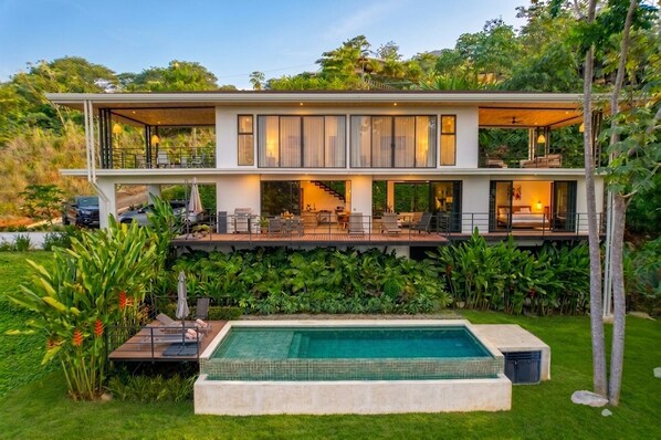 A modern two-story house with a swimming pool, outdoor seating, and lush greenery.