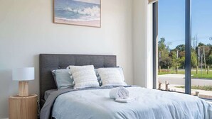 A bedroom with a large window, a bed with grey bedding, a bedside lamp, and a framed picture on the wall.