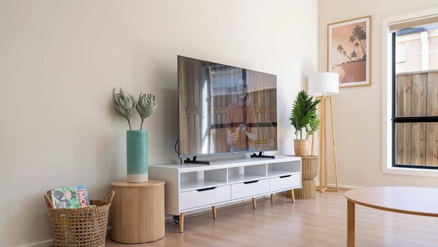 A modern living room with a flat-screen TV, a white entertainment unit, a round wooden table, and a large window.