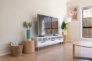 A modern living room with a flat-screen TV, a white entertainment unit, a round wooden table, and a large window.