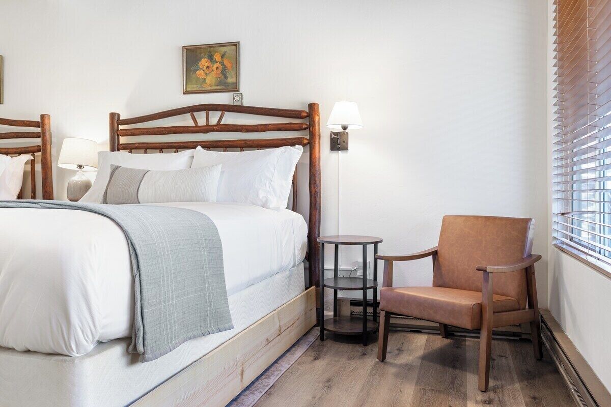 A hotel room with a wooden bed, a chair, a small table, and a window with blinds.