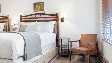 A hotel room with a wooden bed, a chair, a small table, and a window with blinds.