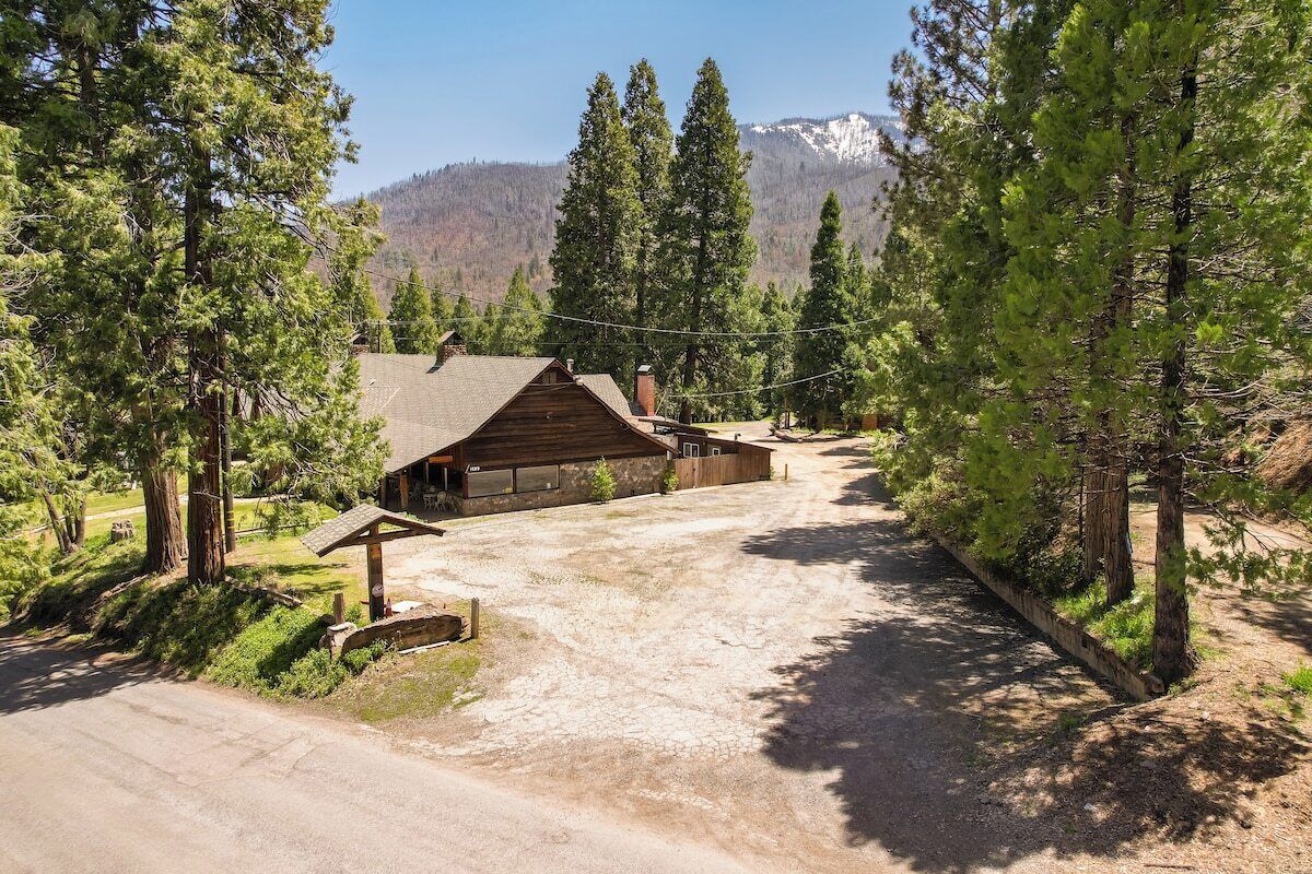 A rustic cabin with a dirt driveway, surrounded by tall pine trees and a mountain backdrop.