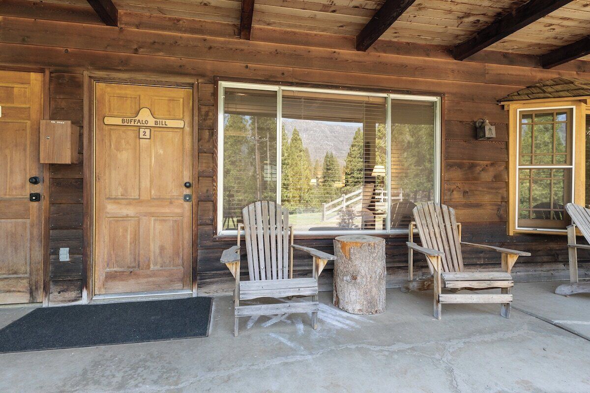 Une cabane en bois avec une porte portant l’inscription « Buffalo Bill », deux fauteuils Adirondack, une table en bois et une grande fenêtre donnant sur une zone montagneuse boisée.