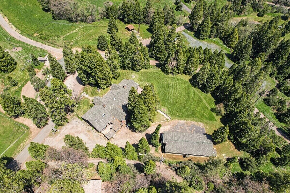 An aerial view of a residential area with multiple houses, surrounded by dense trees and open grassy fields.