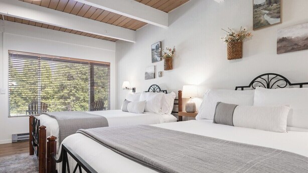 A hotel room with two beds, a large window with blinds, wall-mounted artwork, and a small table with a lamp. - Timberline Retreat Camp Nelson Lodge #3 (Springville)